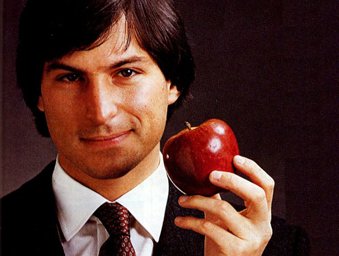 Portrait of a young Steve Jobs, wearing a suit and tie, and holding a red apple in one hand.