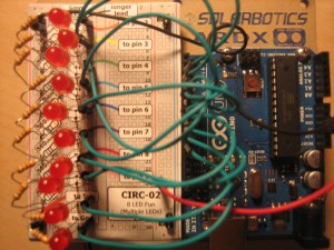 Photo of a breadboard to the left of a blue microchip. There is a column of 10 red LEDs on the breadboard, and they are wired to the chip.